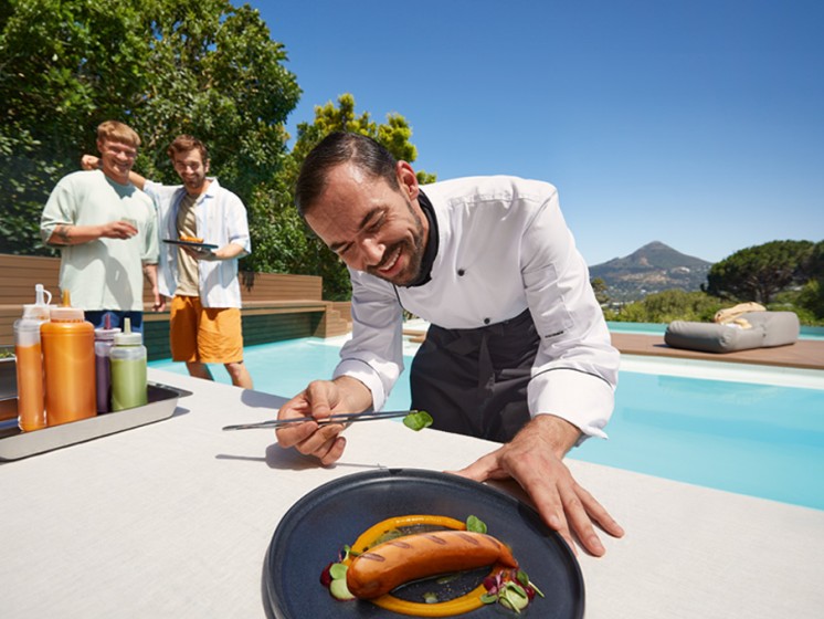 Koch steht vor einem Pool und verziert eine Bratwurst | WestLotto Eurojackpot